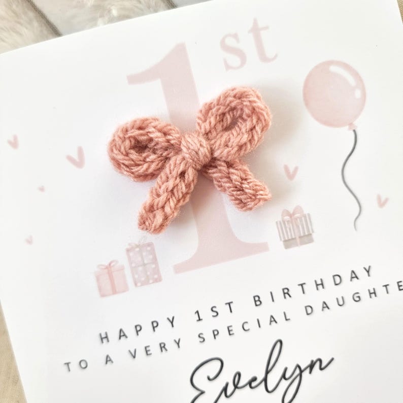 Może przedstawiać: R&oacute;żowa i biała kartka urodzinowa z dzianą kokardką, r&oacute;żowym balonem i tekstem "Happy 1st Birthday to a very special daughter Evelyn".