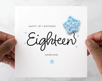 May include: A white card with the text "Happy 18th Birthday" and "Eighteen" in black cursive writing. The name "Harrison" is printed below the cursive writing. A blue crocheted star is attached to the top right corner of the card.