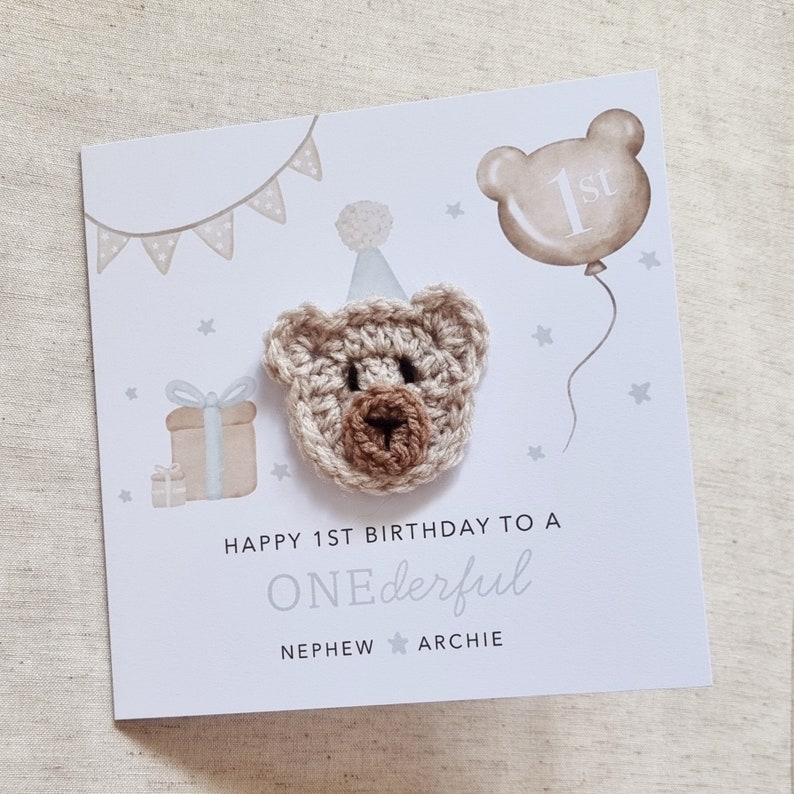 Puede incluir: Una tarjeta blanca con un oso de crochet marr&oacute;n que lleva un gorro de fiesta azul. La tarjeta tiene un globo azul con el n&uacute;mero "1er" y el texto "Feliz 1er cumplea&ntilde;os a un SOBRINO ONEderful ARCHIE".