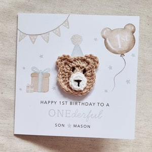 Puede incluir: Una tarjeta blanca con un oso de crochet marr&oacute;n que lleva un gorro de fiesta. La tarjeta dice "Happy 1st Birthday to a ONEderful SON MASON".