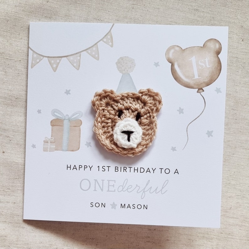 Puede incluir: Una tarjeta blanca con un oso de crochet marr&oacute;n que lleva un gorro de fiesta. La tarjeta dice "Happy 1st Birthday to a ONEderful SON MASON".
