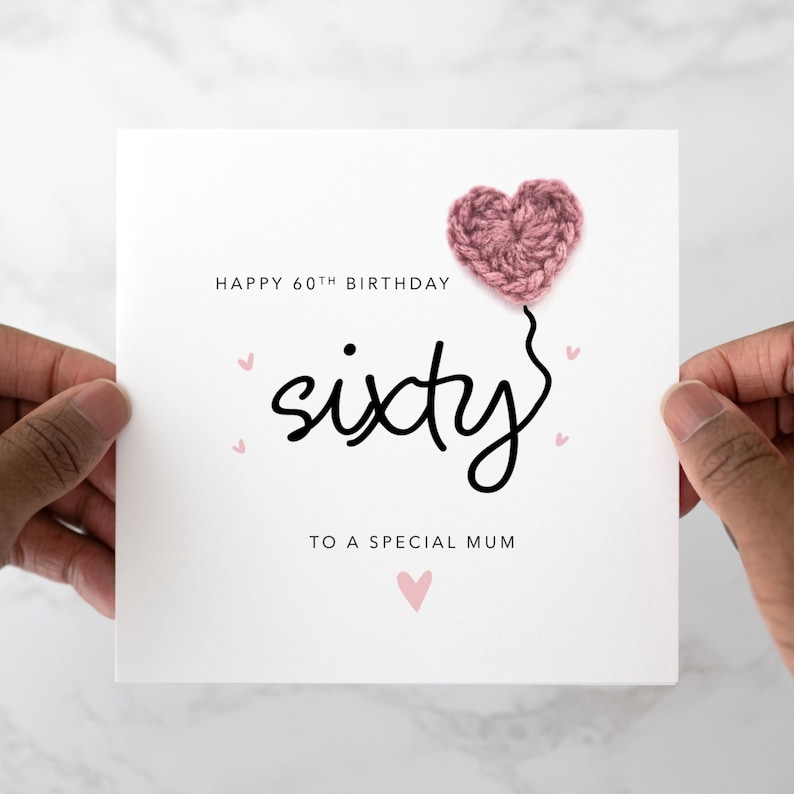 Peut inclure: Une carte blanche avec le texte "Happy 60th Birthday sixty to a special Mum" et un ballon en forme de c&oelig;ur en crochet rose.