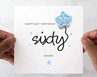 May include: A white card with the text "Happy 60th Birthday" and "sixty" in black script. A blue crocheted star is attached to the card. The name "Jason" is written below the text.