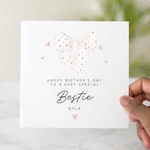 Può includere: Un biglietto di auguri bianco per la festa della mamma con un fiocco ad acquerello e cuori rosa. Il biglietto recita "Happy Mother's Day to a Very Special Bestie" con il nome "Nyla" sotto. Una mano tiene il biglietto.