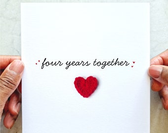 May include: A white card with the text "four years together" in black script, accompanied by a small red crocheted heart. The card is held by hands, suggesting a gift or greeting card.