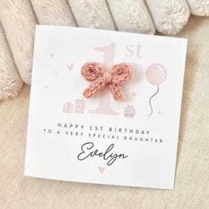 Może przedstawiać: R&oacute;żowa i biała kartka urodzinowa z r&oacute;żową dzianą kokardką z przodu. Na kartce napisano "Happy 1st Birthday to a very special daughter Evelyn".