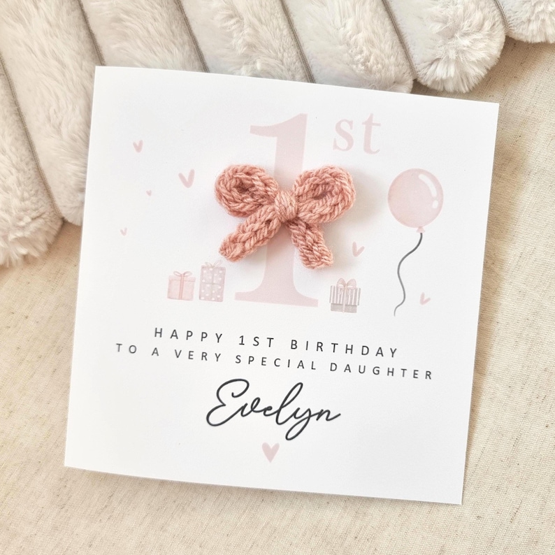 Może przedstawiać: R&oacute;żowa i biała kartka urodzinowa z r&oacute;żową dzianą kokardką z przodu. Na kartce napisano "Happy 1st Birthday to a very special daughter Evelyn".