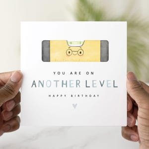 May include: A white greeting card with a yellow level tool illustration with a face. The text reads "YOU ARE ON ANOTHER LEVEL HAPPY BIRTHDAY". The card is held by two hands.