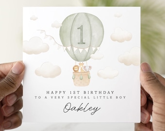 May include: A white card with a green hot air balloon with a number 1 on it. The balloon is surrounded by white clouds and has three animals in the basket. The text on the card reads "Happy 1st Birthday to a very special little boy Oakley".