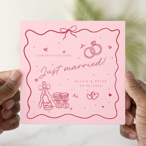 May include: A pink wedding card with red line art. The card says "Just married!" with the names Olivia & Peter and the date 28.06.2026. The card has illustrations of rings, champagne, and hearts.