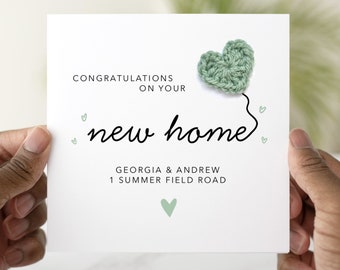 Puede incluir: Una tarjeta blanca con un corazón de crochet verde sujeto a la parte superior. La tarjeta dice "Felicidades por su nuevo hogar" con los nombres Georgia & Andrew y la dirección 1 Summer Field Road.