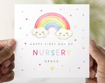 May include: A white card with a watercolor rainbow and smiling clouds. The card reads "Happy First Day of Nursery" and the name "Grace" in colorful letters. Small colorful dots and a yellow heart decorate the card.