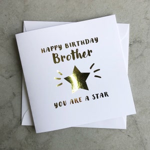 Può includere: Un biglietto bianco con lettere dorate che recitano "Buon compleanno Fratello Sei una stella". Una stella dorata è al centro del biglietto.