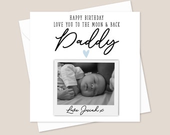 May include: A black and white photo of a baby sleeping in a white onesie. The photo is in a white frame with a blue heart above it. The text "Happy Birthday Love you to the moon & back Daddy" is above the photo and the text "Love Josiah x" is below the photo.