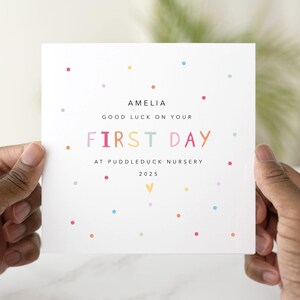 May include: A white card with colourful polka dots and text. The text reads "AMELIA GOOD LUCK ON YOUR FIRST DAY AT PUDDLEDUCK NURSERY 2025". The card is held by two hands.