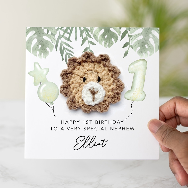 May include: A white card with green leaves printed on it. The card has a crocheted lion head, a green balloon with a star, and a green number one balloon. The card says "Happy 1st Birthday to a very special nephew Elliot".