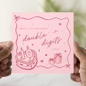 May include: A pink birthday card with red line art. The card reads "Happy 10th Birthday" and "double digits" with a cake, gifts, and hearts. The name "Lola" is also on the card.