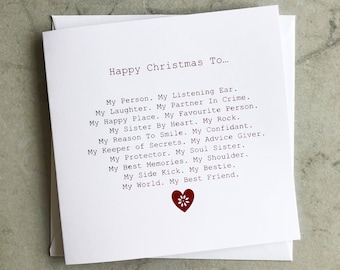 Può includere: Un biglietto bianco con testo rosso che recita "Buon Natale a..." seguito da un elenco di frasi affettuose che descrivono una relazione stretta. Un cuore rosso con un disegno di fiore bianco si trova nella parte inferiore del biglietto.
