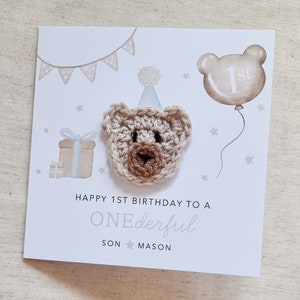 Puede incluir: Una tarjeta blanca con un oso de crochet marr&oacute;n que lleva un gorro de fiesta azul. La tarjeta dice "Happy 1st Birthday to a ONEderful SON MASON". Hay una caja de regalo azul y blanca, un globo con el n&uacute;mero "1st" y estrellas en el fondo.
