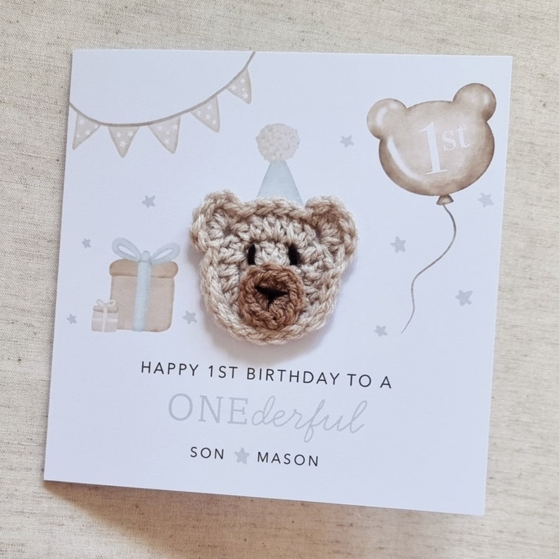 Puede incluir: Una tarjeta blanca con un oso de crochet marr&oacute;n que lleva un gorro de fiesta azul. La tarjeta dice "Happy 1st Birthday to a ONEderful SON MASON". Hay una caja de regalo azul y blanca, un globo con el n&uacute;mero "1st" y estrellas en el fondo.