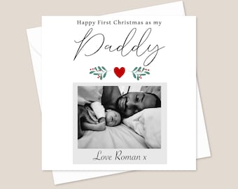 May include: A white Christmas card with the text "Happy First Christmas as my Daddy" in black script. A black and white photo of a father and baby is below the text. The card includes a red heart and green sprigs.