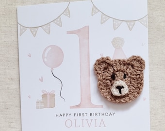 May include: A pink and white birthday card with a large pink number one, a brown crocheted bear wearing a party hat, a pink balloon, and a gift box. The card says "Happy First Birthday Olivia".