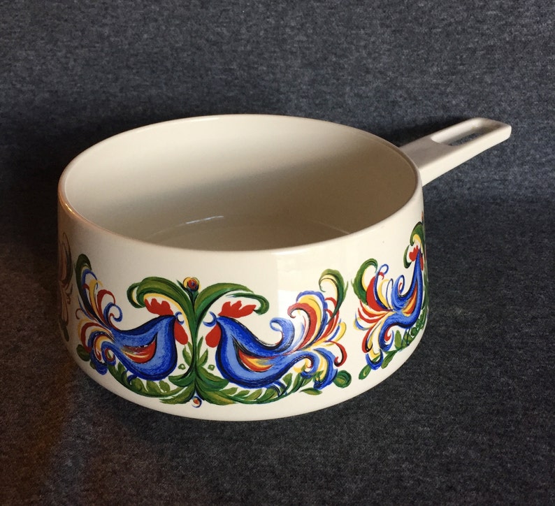 May include: White ceramic pot with a blue, red, yellow, and green floral design featuring stylized birds. The pot has a white plastic handle.