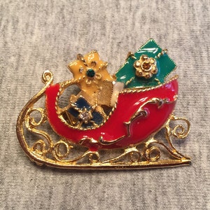 May include: A festive Christmas brooch featuring a red sleigh with gold filigree detailing. The sleigh is filled with wrapped presents in yellow, green, and blue, adorned with sparkling gemstones. A charming holiday accessory.