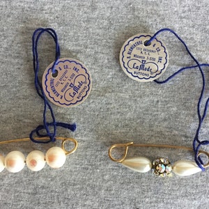 May include: Two gold safety pins with pearl and rhinestone accents. Each pin is attached to a round tag with the text "La Mode Buttons" and the company name and address.
