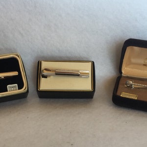 May include: Three tie bars in their original boxes. The tie bars are gold, silver, and a combination of gold and silver. The boxes are black velvet with gold trim.
