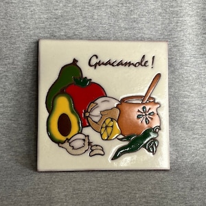 May include: A white ceramic tile with a colorful illustration of guacamole ingredients, including an avocado, tomato, garlic, lime, and a bowl with a spoon. The text "Guacamole!" is written in brown cursive.