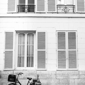 May include: Black and white photograph of a vintage bicycle parked in front of a building with white walls and shutters. The bicycle has a basket on the front and a bag on the back. The building has multiple windows.