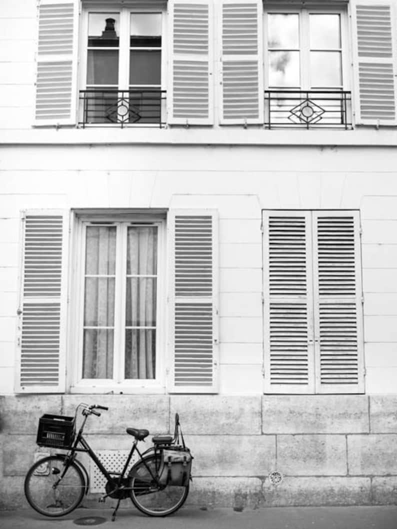 May include: Black and white photograph of a vintage bicycle parked in front of a building with white walls and shutters. The bicycle has a basket on the front and a bag on the back. The building has multiple windows.