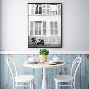 May include: Black and white framed print of a Parisian street scene. A bicycle is parked in front of a building with shutters. A small round table with two white chairs, a plant, and tableware is in the foreground.