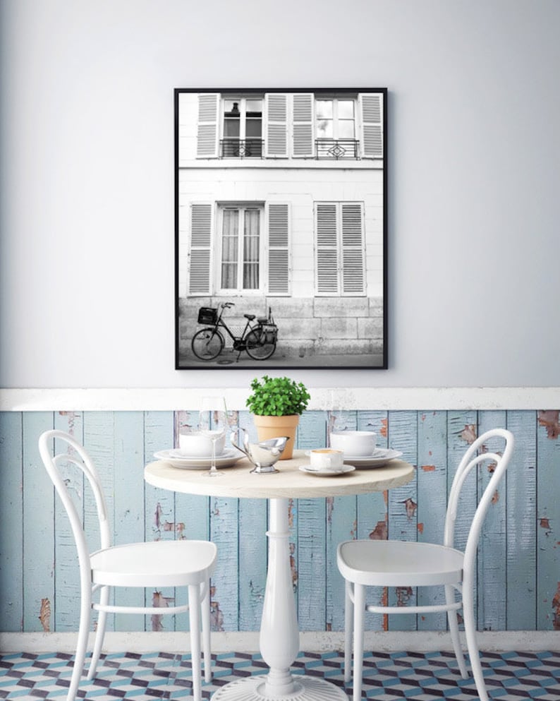 May include: Black and white framed print of a Parisian street scene. A bicycle is parked in front of a building with shutters. A small round table with two white chairs, a plant, and tableware is in the foreground.