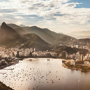 May include: An aerial view of a city with a large body of water in the foreground. The city is surrounded by mountains, and there are many boats in the water. The sky is blue and cloudy.