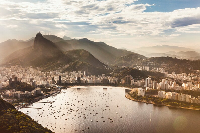 May include: An aerial view of a city with a large body of water in the foreground. The city is surrounded by mountains, and there are many boats in the water. The sky is blue and cloudy.