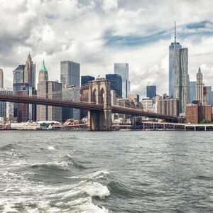 Puede incluir: Una vista del puente de Brooklyn con el horizonte de la ciudad de Nueva York al fondo. El puente est&aacute; hecho de acero y tiene un color marr&oacute;n. El agua est&aacute; agitada y gris.