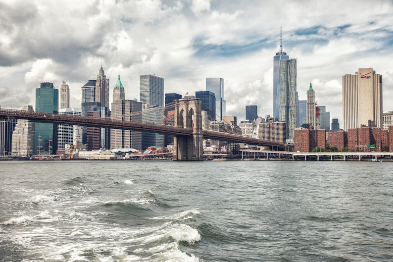 Puede incluir: Una vista del puente de Brooklyn con el horizonte de la ciudad de Nueva York al fondo. El puente est&aacute; hecho de acero y tiene un color marr&oacute;n. El agua est&aacute; agitada y gris.
