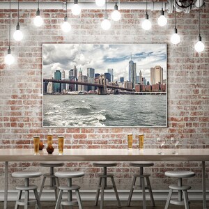 Puede incluir: Una pared de ladrillo con tuber&iacute;as expuestas y bombillas colgantes sobre una mesa de bar con taburetes. Una gran impresi&oacute;n enmarcada del horizonte de la ciudad de Nueva York con el puente de Brooklyn cuelga de la pared.