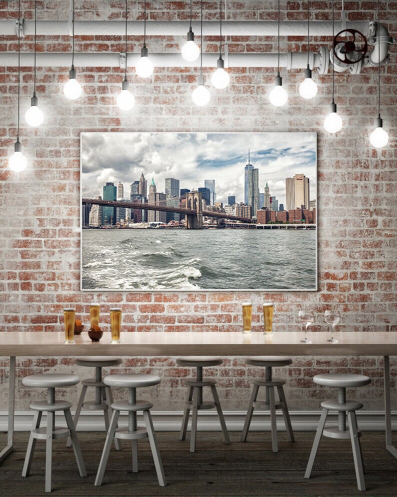 Puede incluir: Una pared de ladrillo con tuber&iacute;as expuestas y bombillas colgantes sobre una mesa de bar con taburetes. Una gran impresi&oacute;n enmarcada del horizonte de la ciudad de Nueva York con el puente de Brooklyn cuelga de la pared.