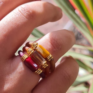 May include: A set of three stacked rings featuring different coloured stones. The top ring has a yellow stone, the middle ring has a brown stone, and the bottom ring has a red stone. The rings are made of gold and have a simple design.
