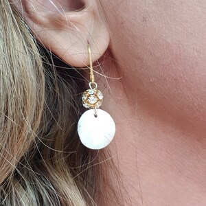 Puede incluir: Pendientes de aro dorados con un disco redondo blanco y una pequeña bola dorada con brillantes transparentes.