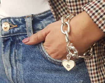 May include: A silver chain bracelet with a heart charm featuring a small flower design. The bracelet is worn on a person's wrist over a blue denim jean.