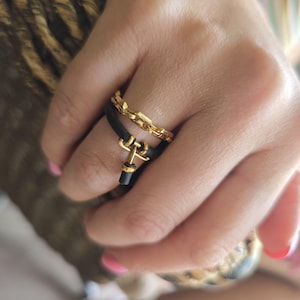 May include: A close-up of two rings on a finger. One is a gold chain ring, and the other is a black band with a gold anchor-like charm. The rings are worn on a hand with pink nail polish.