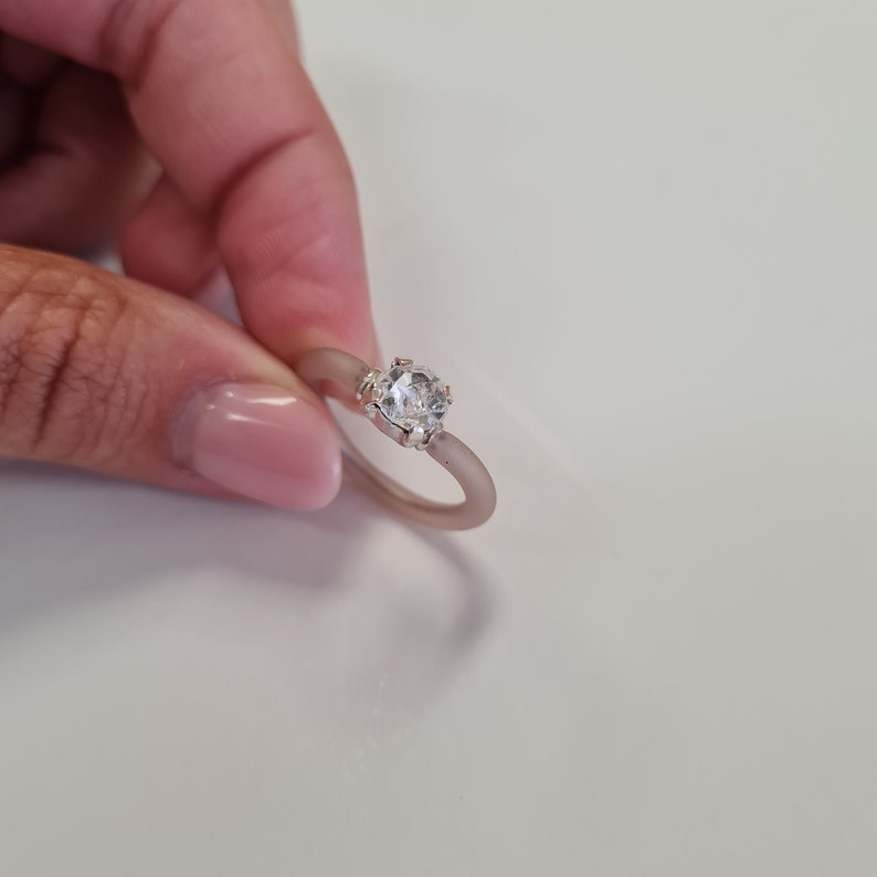 K&ouml;nnte beinhalten: Ein silberner Ring mit einem gro&szlig;en, klaren, runden Edelstein. Das Ringband hat eine helle, ged&auml;mpfte rosa Farbe. Der Edelstein wird von einer silbernen Fassung gehalten.