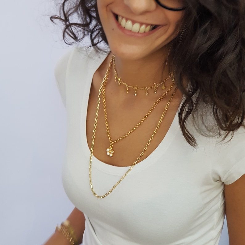 May include: A woman wearing a white t-shirt with a gold layered necklace featuring a small flower pendant and a delicate chain with star and circle charms.
