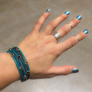 Puede incluir: Una mano con un brazalete de múltiples capas con correas de cuero turquesa y marrón y detalles plateados. El brazalete tiene pequeñas piedras turquesas. La mano también lleva varios anillos plateados.