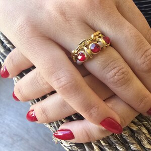 May include: A gold-toned ring with three red gemstones. The ring is on a hand with red nail polish. The ring has a unique design with a chain-like pattern. The gemstones are round and set in a bezel setting.