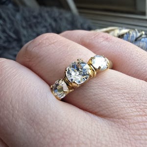 Op de afbeelding: Een gouden ring met drie heldere, gefacetteerde edelstenen. De centrale steen is de grootste, geflankeerd door twee kleinere stenen. De band van de ring is van gepolijst goud. De ring wordt aan een vinger getoond.
